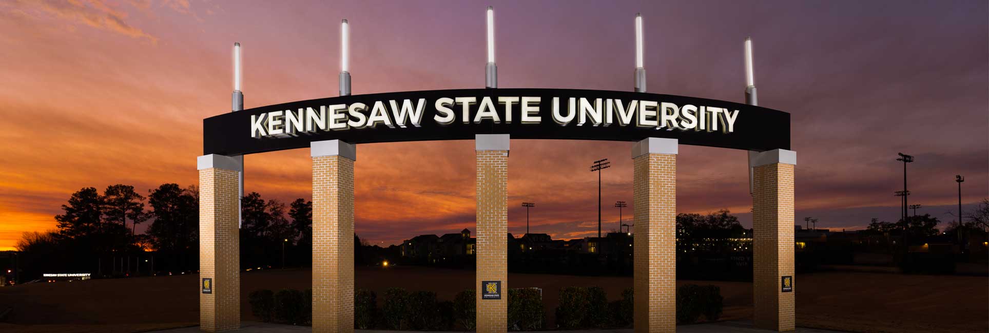 An illuminated Kennesaw State University entrance sign arches across six brick columns, glowing against a vivid sunset sky with rich hues of orange, purple, and pink.