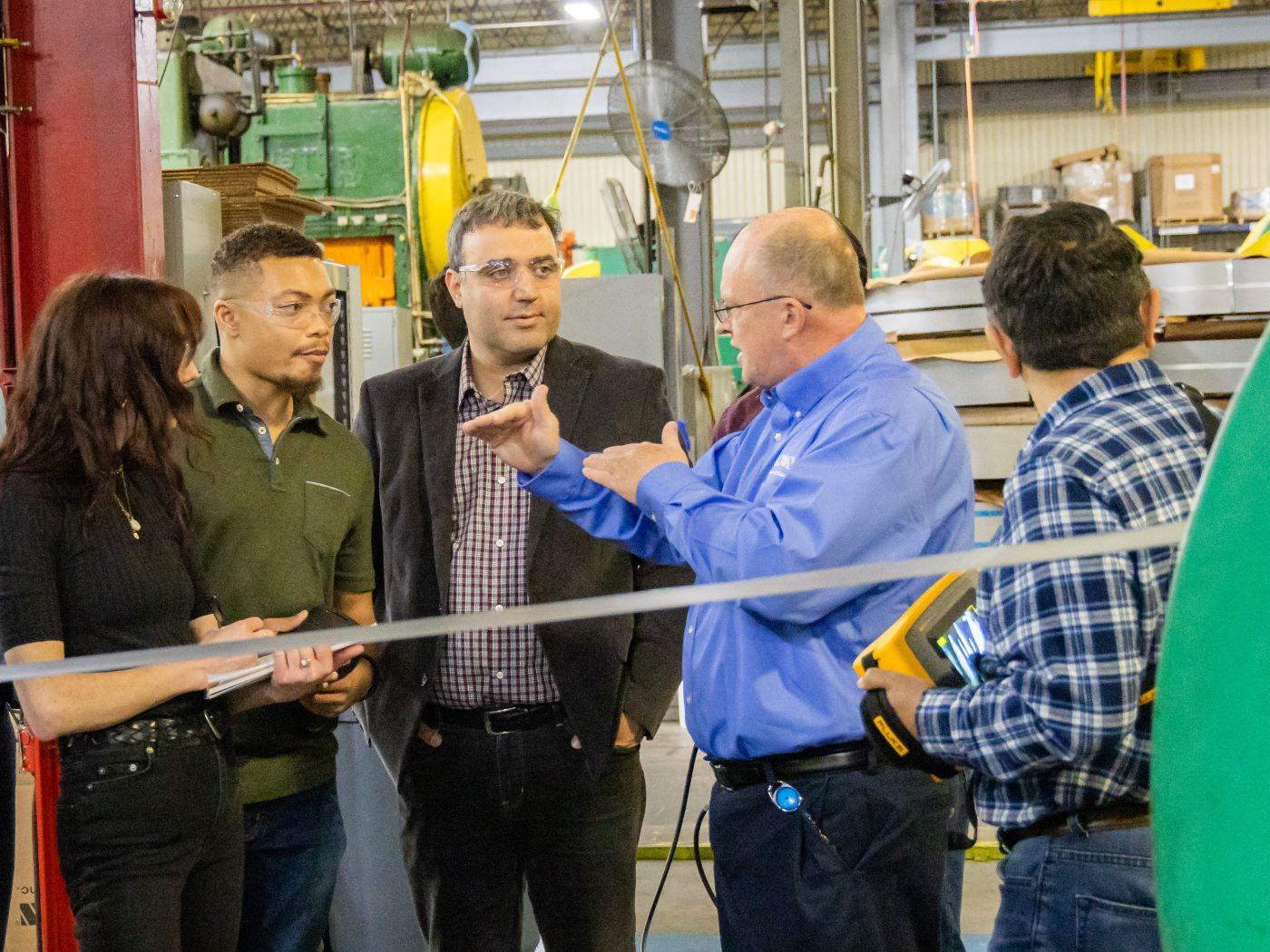 Five people standing in an industrial setting, engaged in discussion. / 