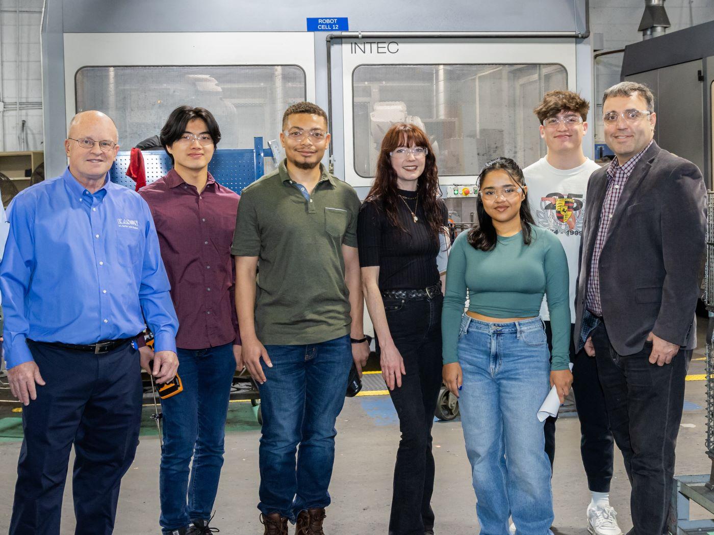 Seven people standing in front of industrial machinery in a factory. / 