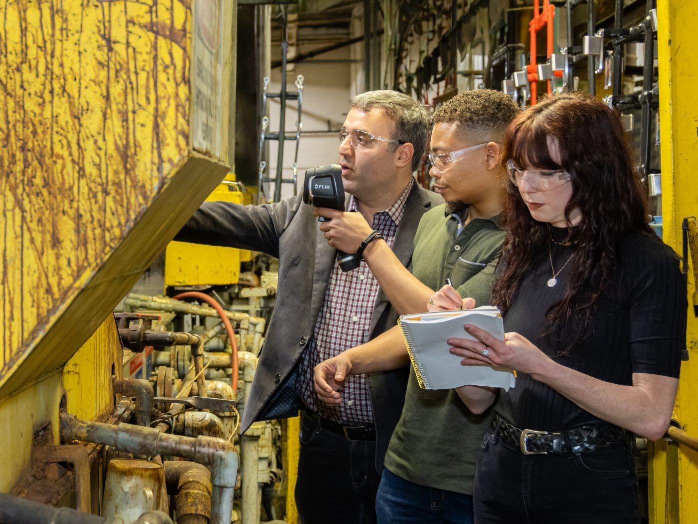Three people inspecting yellow industrial machinery, one holding a device and another taking notes. / 
