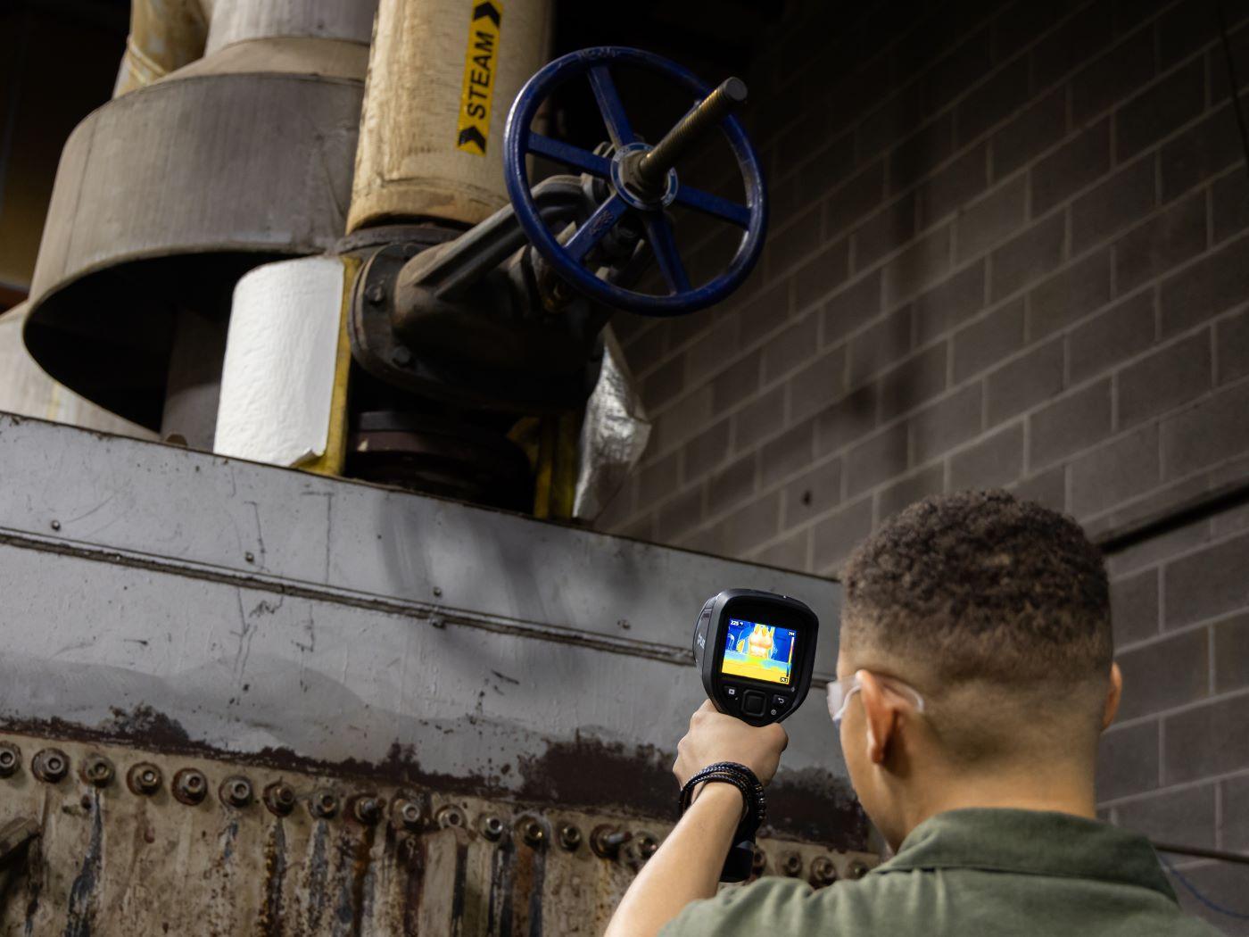 Person using a thermal imaging camera to inspect a pipe with a blue valve labeled "STEAM." / 