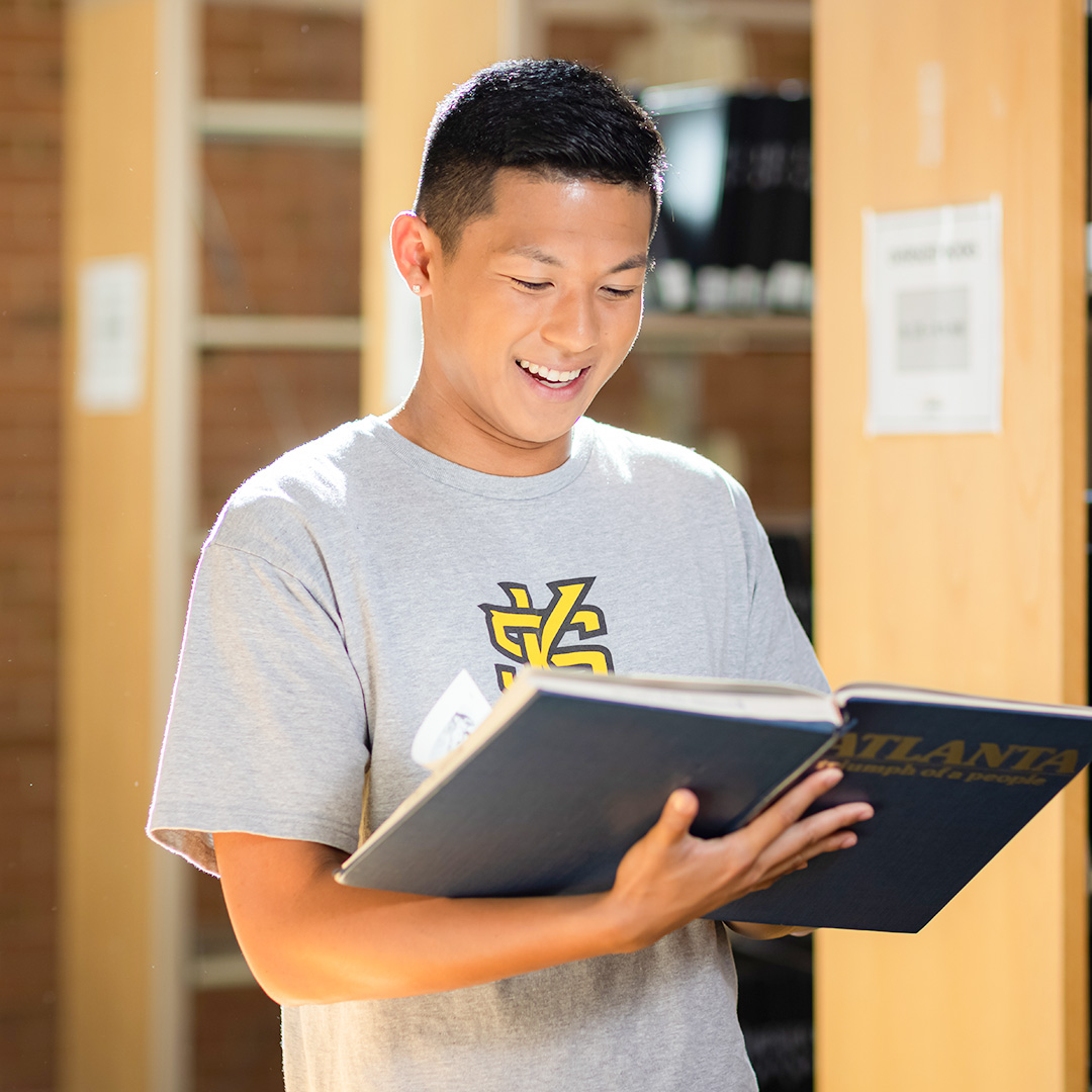 male student with gray ksu shirt opening a book and smiling in library