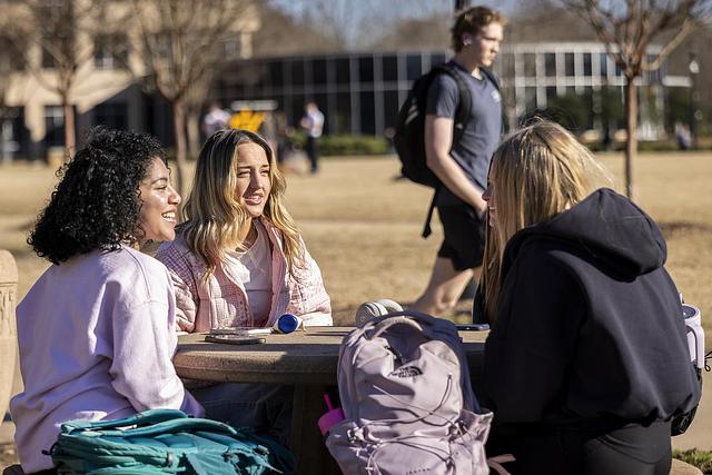 Three young women sit at a picnic table, chatting and laughing, with backpacks around them. A person with a backpack walks by in the sunny ksu setting.