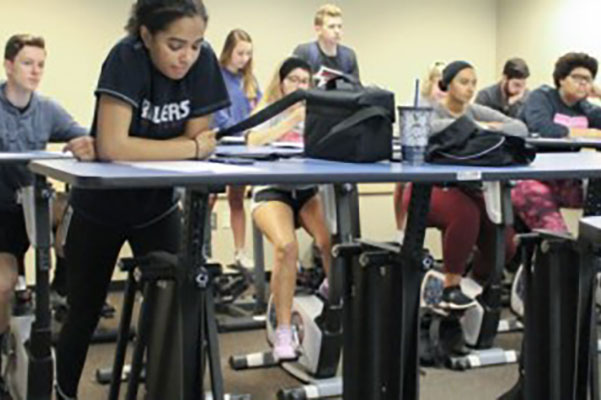 students in class on stationary bikes with desks. 
