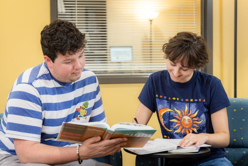Two students sitting next to each other, one holding a book and the other writing in a notebook.