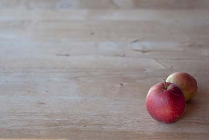 Two red apples are clustered in one corner of a wooden table, drawing attention to the fruit in an open space. Image is titled “apples” by Paul Bausch