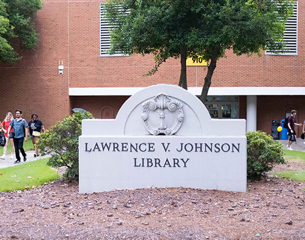 A white stone sign reads "Lawrence V. Johnson Library". The sign is located outdoors in a landscaped area with mulch and shrubbery, in front of a large red brick building.
