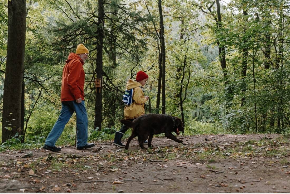 old man and young boy in jackets with black dog walking thru the woods