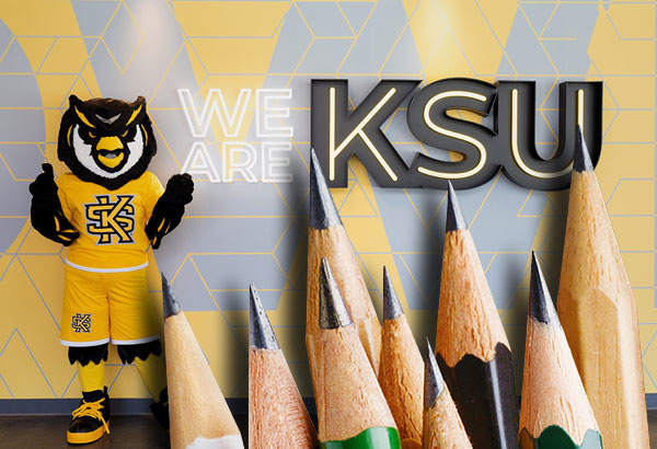 Kennesaw State University mascot standing next to a wall with the words 'We Are KSU' and a group of sharpened pencils in the foreground.